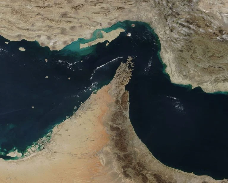 Satellite view of a desert peninsula extending into dark blue sea with turquoise shallow waters along the coast, showing arid terrain.