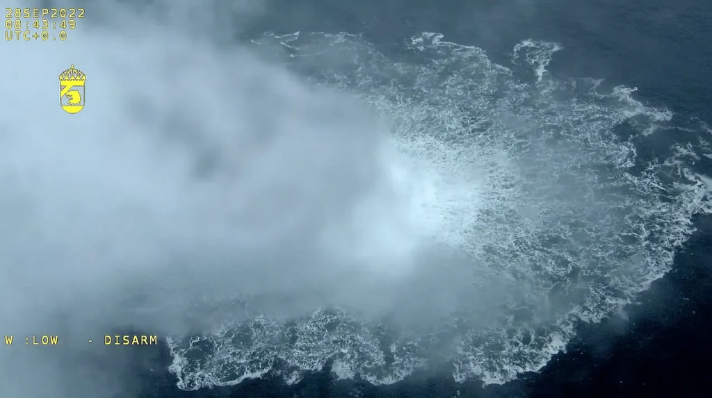 Drone footage of a controlled detonation over the ocean creating a white spray ring; timestamp and 'DISARM' text visible.