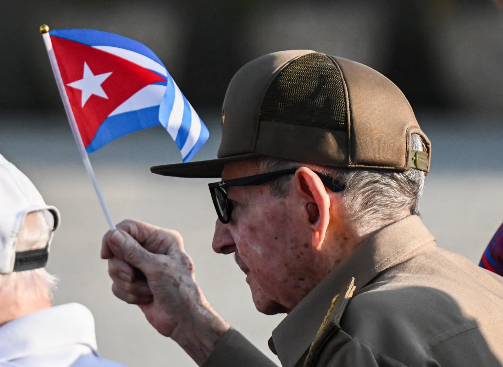Older man wearing a brown cap and sunglasses holds a small Cuban flag outdoors, engaging with someone off-camera.] ,