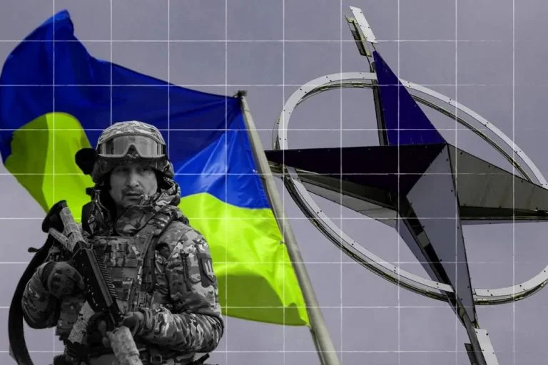 Soldier in camouflage with a rifle in front of a waving blue-and-yellow Ukrainian flag against a gray sky.